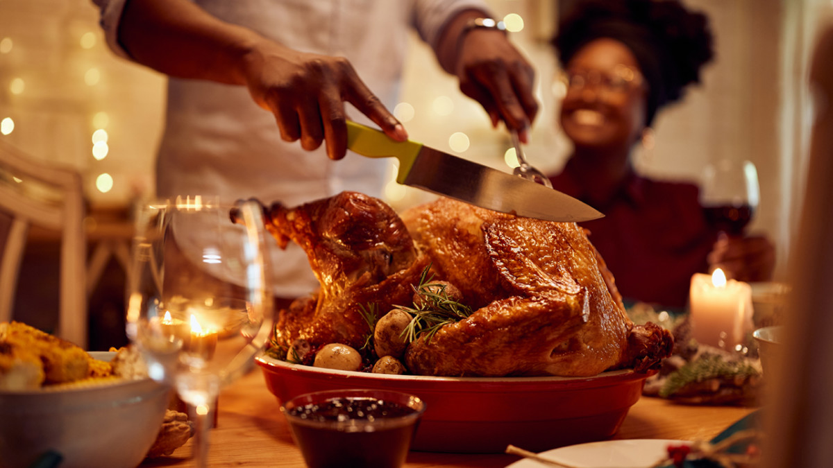 Close-up of black man carving roasted turkey during Thanksgiving meal at dining table.