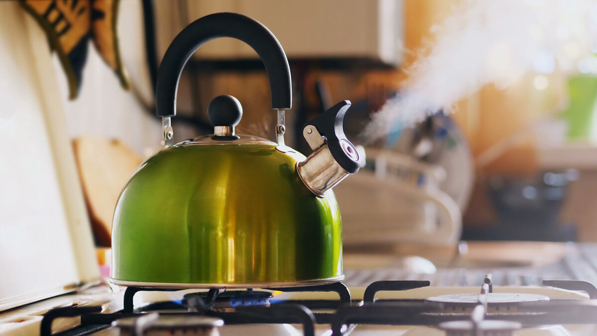 Kettle boiling on a gas stove. Boiling green kettle boiling with steam emitted from spout. Shallow depth of field. Solar glare from the kitchen window